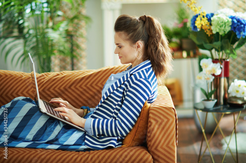 woman laying on sofa with laptop in modern house in sunny day