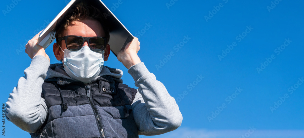 A young man in sunglasses and medical mask against blue sky covers his head from sun and viruses with modern laptop. Working remotely during coronavirus pandemic. Spring 2020. Man hiding under laptop A young man in sunglasses and medical mask against blue sky covers his head from sun and viruses with modern laptop. Working remotely during coronavirus pandemic. Spring 2020. Man hiding under laptop