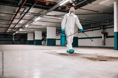 Worker in sterile uniform and mask walking trough underground garage and sterilizing surface. Protection from corona virus / covid-19 concept. © dusanpetkovic1