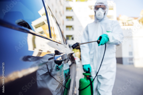 Worker in sterile uniform and mask standing outdoors and sterilizing car from corona virus / covid-19. Selective focus on sprayer.
