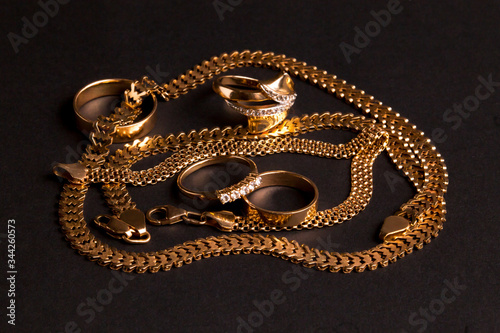 Various gold jewelry, chaotically arranged: wedding rings, engagement ring, chain, bracelet. Isolated on a matte black background.