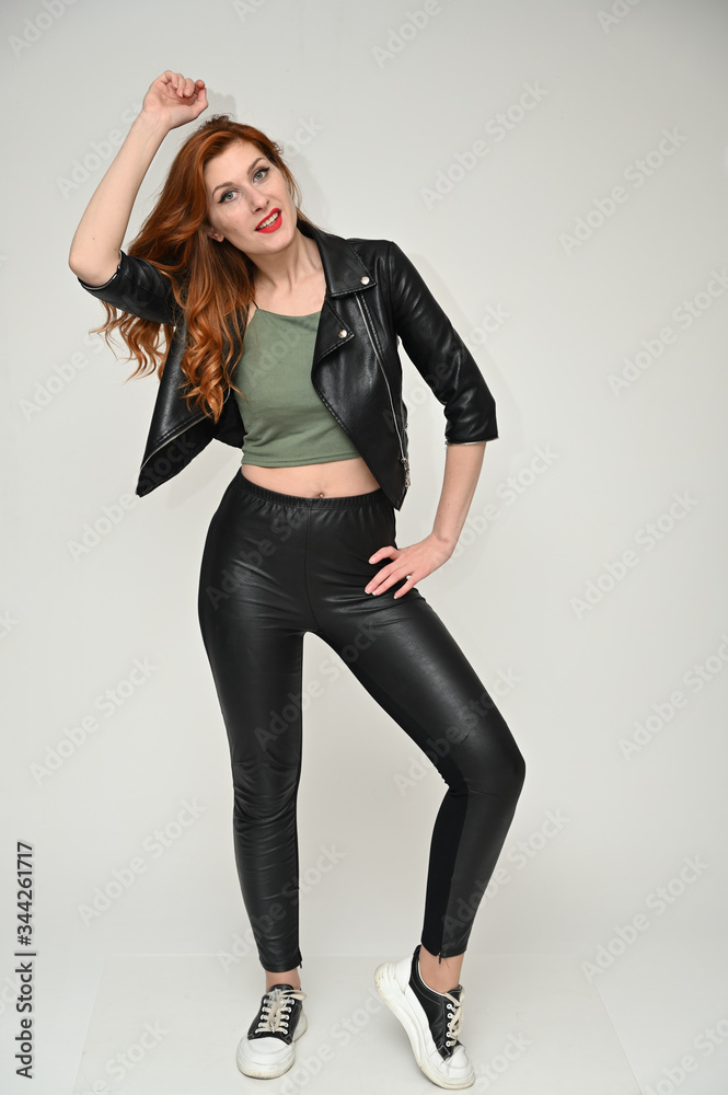 Vertical portrait on a white background of a pretty red-haired young woman. A full-length model with excellent makeup poses in a black jacket and trousers in the studio. Vertical portrait on a white background of a pretty red-haired young woman. A full-length model with excellent makeup poses in a black jacket and trousers in the studio.