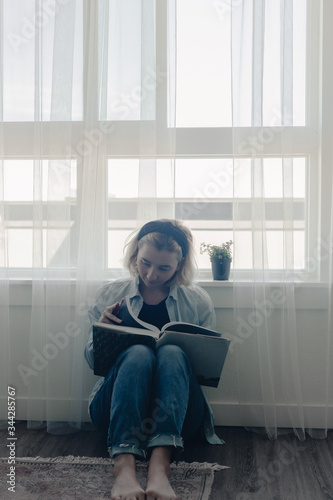 Wallpaper Mural Lady sits in front of large window on the floor. Social distancing. Self isolation, stay at home. Dreaming of future. Spring time at home. Reading book near window. White curtains. Reading fiction. Torontodigital.ca