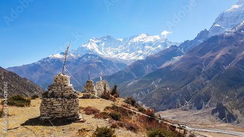 A row of small stony stupas with Annapurna Chain as a backdrop, Himalayas, Nepal. High mountains covered with snow. Land in front of the stupa is barren and dry. Some prayer's flag next to it.