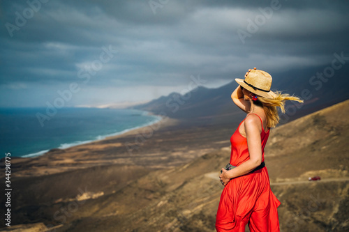 Beautiful young woman with red vibrant dress looking at rocky mountains and beach of Cofete in Fuerteventura