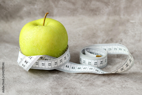 A ripe green fresh Apple wrapped in white measuring tape lies on a gray background. Diet and weight loss. Vegetarianism. Selective focus
