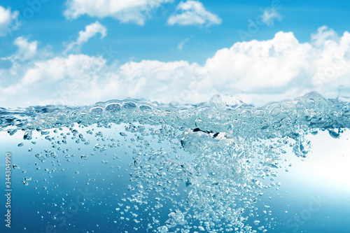 Blue water wave with splash and bubbles surface on blue sky with white clouds