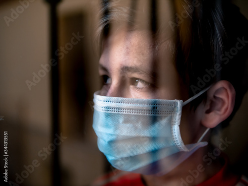 Boy with a medical mask looks sadly out of the window during his confinement because of convid 19