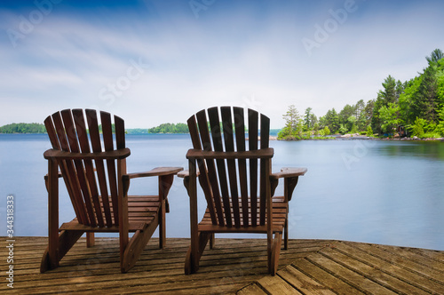 Two Muskoka chairs sitting on a wood dock facing a lake. Across the calm water is a white cottage nestled among green trees. There is a boat dock on the water in front of the cottage.