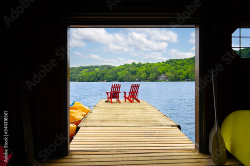 View of two red Adirondack chair on a wooden dock from a cottage's boathouse in Muskoka, Ontario Canada. In the background cottages are nestled between trees. photo