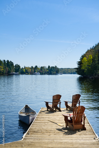 Fotografia Three Adirondack chairs on a wooden dock on a calm lake in Muskoka, Ontario Canada