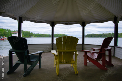 Three colourful Adirondack chairs overlooking a calm lake under a gazebo. Cottages nestled between green trees are visible across the water. The chairs are green  yellow and red.
