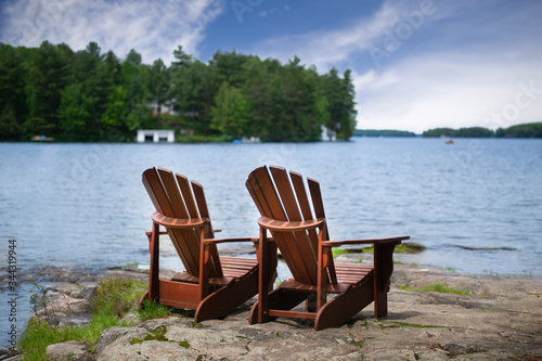 Two Muskoka chairs along the shore facing a lake in Ontario, Canada. A white cottage is visible across the blue water nestled between green trees.