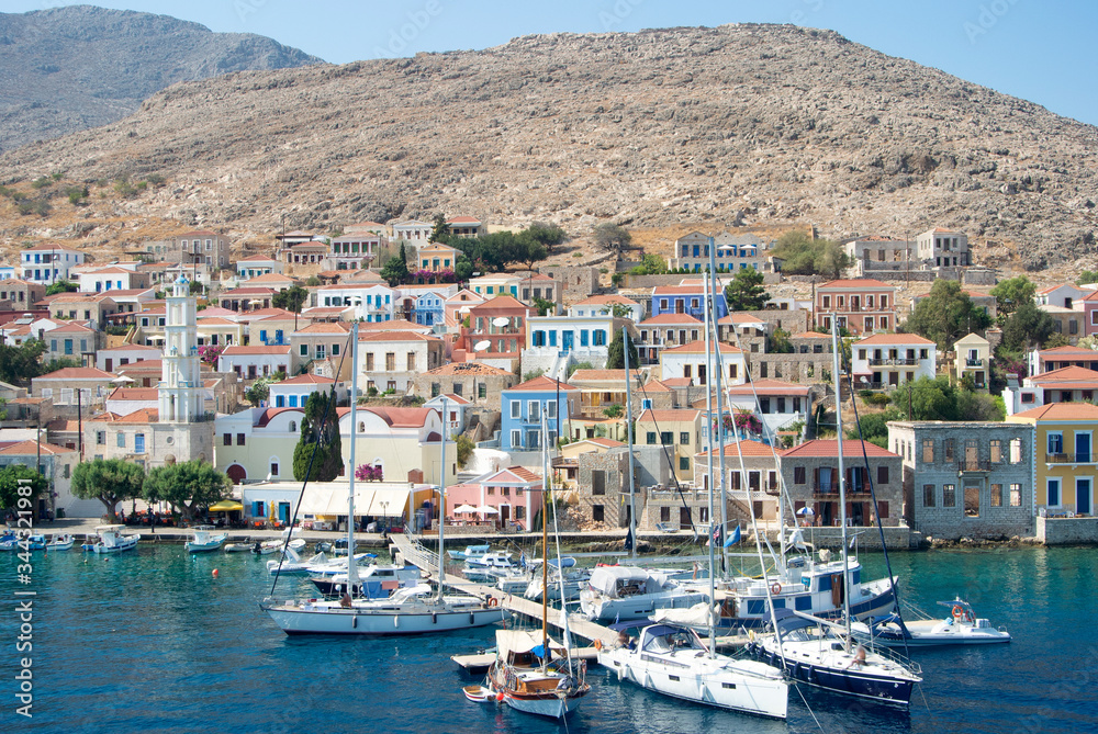 The colourful harbour at the the beautiful Greek island of Chalki. Classical architecture rises from the waterfront looking over the bay. A warm summers day with pleasure boats moored. The colourful harbour at the the beautiful Greek island of Chalki. Classical architecture rises from the waterfront looking over the bay. A warm summers day with pleasure boats moored.