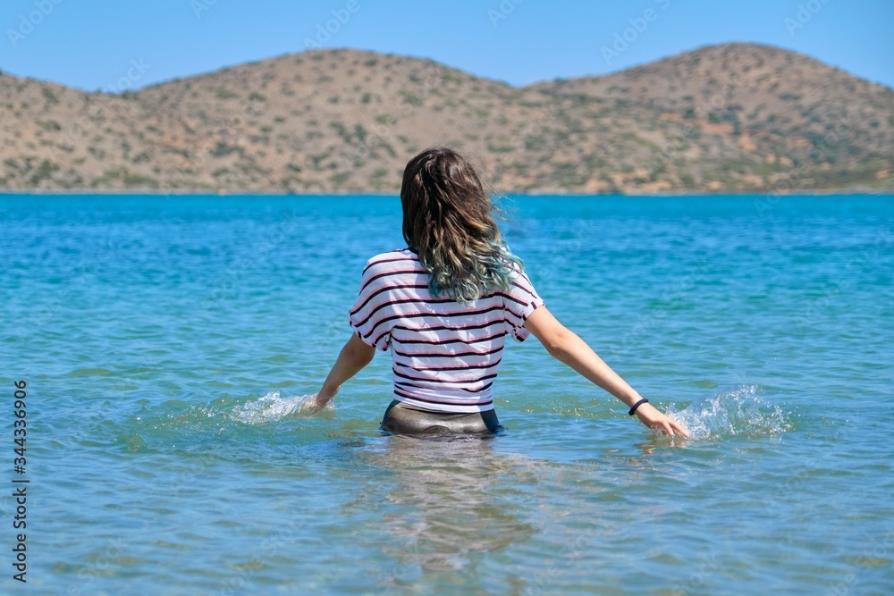 Teenager girl is touching, enjoying sea water in the bay, back view Teenager girl is touching, enjoying sea water in the bay, back view