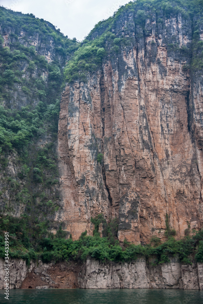 Wuchan, China - May 7, 2010: Dragon Gate Gorge on Daning River. Tall brown and black cliff descending into emerald green water. Green foliage on top and bottom. Silver sky..