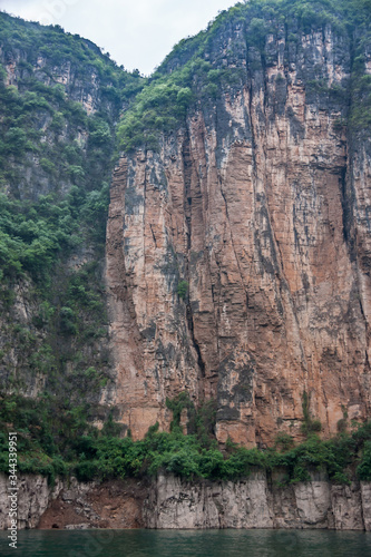 Wuchan, China - May 7, 2010: Dragon Gate Gorge on Daning River. Tall brown and black cliff descending into emerald green water. Green foliage on top and bottom. Silver sky.. © Klodien