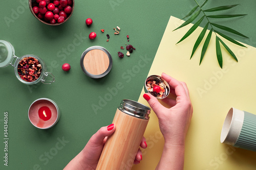  Trendy creative flat lay with hands, two tone green and yellow paper. Zero waste tea to go, making herbal infusion in eco friendly insulated bamboo steel flask with herbal mixture and cranberry. photo