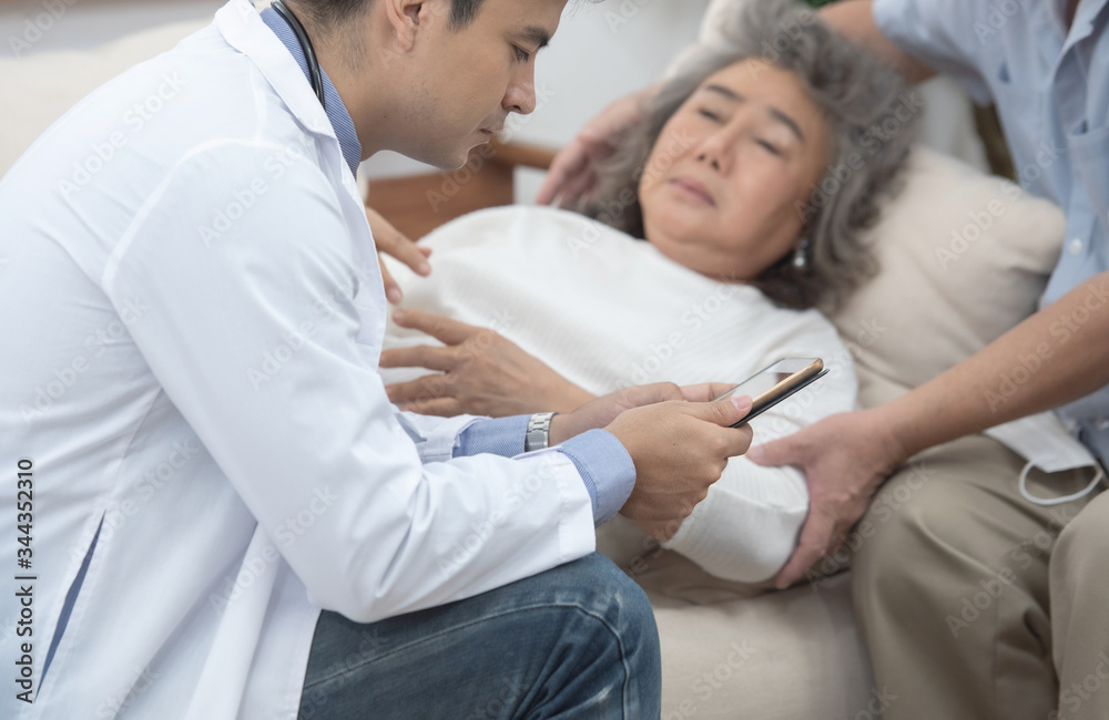 Fototapeta premium Asian senior elderly old patient woman laying down on sofa in home waiting for result from smart young caucasian doctor smartphone while check up in home with grandfather take care beside.
