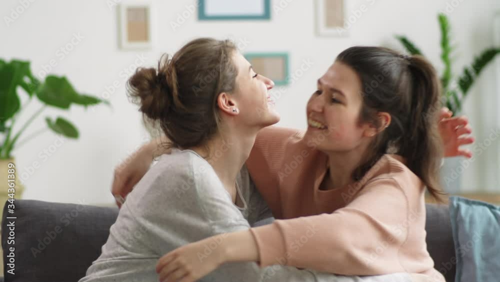 Two young Caucasian women taking medical face masks off, embracing each other and happily smiling while sitting together on couch at home and celebrating the end of coronavirus lockdown Two young Caucasian women taking medical face masks off, embracing each other and happily smiling while sitting together on couch at home and celebrating the end of coronavirus lockdown
