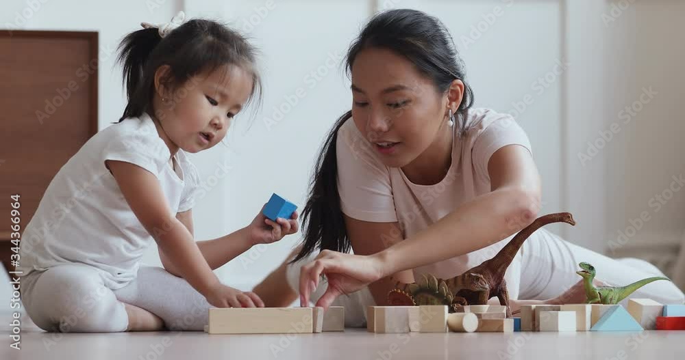 Excited young vietnamese nanny babysitter lying on heated floor with cute preschool child girl, playing with favorite toys at home. Happy asian mommy enjoying free leisure time with baby daughter.