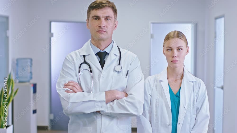 Main doctor and his nurse assistant looking straight to the camera they stand in the middle of the hospital corridor have a serious face Main doctor and his nurse assistant looking straight to the camera they stand in the middle of the hospital corridor have a serious face
