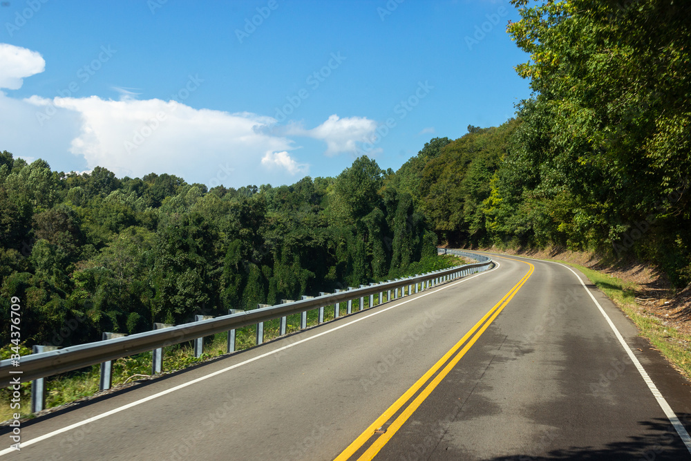 Asphalt road along the trees.  Beautiful empty highway with yellow lines between green trees on a summer warm day with blue sky and fluffy clouds