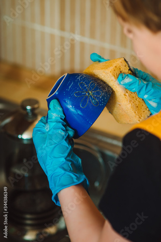 The boy in a blue rubber glove holds a sponge in his hands. The boy helps his mother in the kitchen with cleaning dirty dishes. Mom's assistant. Wash the cup