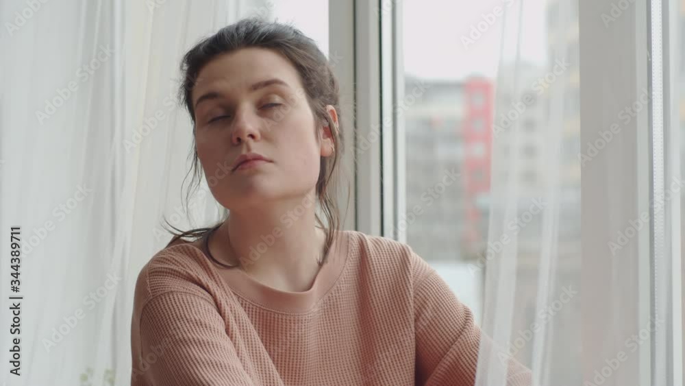 Young sad woman sitting on sill and looking through the window while getting bored at home during self isolation time
