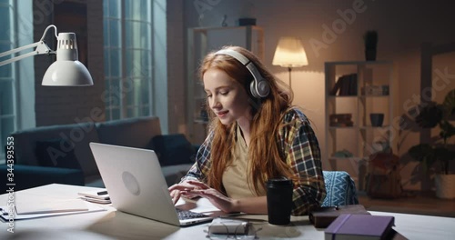 Positive redhead caucasian high school or university student listening to music in headphones and concetrating on work. Young freelancer enjoying remote job in laptop 4k footage photo