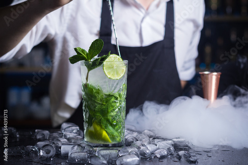 Glass of mojito dressed up with lemon and mint. Ice cubes placed on surface around mojito drink. Little white cloud of smoke hiding measuring cup.Barmen in white top and black apron stirring cocktail