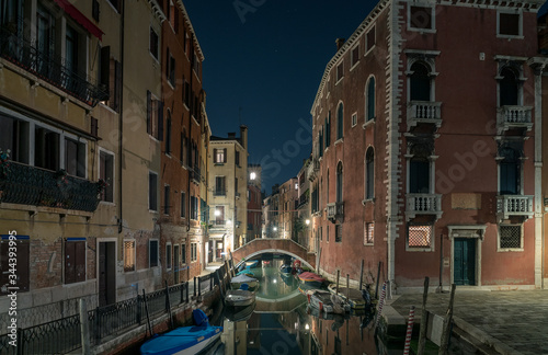 A scenic view of a beautiful Venetian canal at night with colourful architecture, a bridge and reflections of boats running along the water in the town of Venice, Italy © Matthew