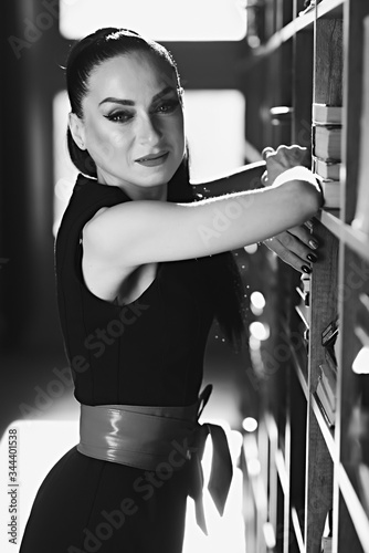 A beautiful brunette girl, a model, with a tattoo in the studio. sun and shadow. Stands in a sexy dress with a red banter of a closet wax with books. looks aside. the photo is black white