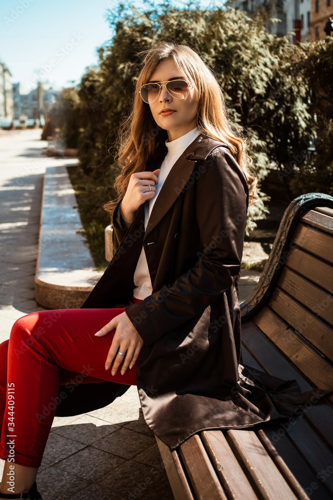Outdoor fashion details. Young brunette woman in coat sitting on a bench in the city. Sunny spring day. Outdoor fashion details. Young brunette woman in coat sitting on a bench in the city. Sunny spring day.