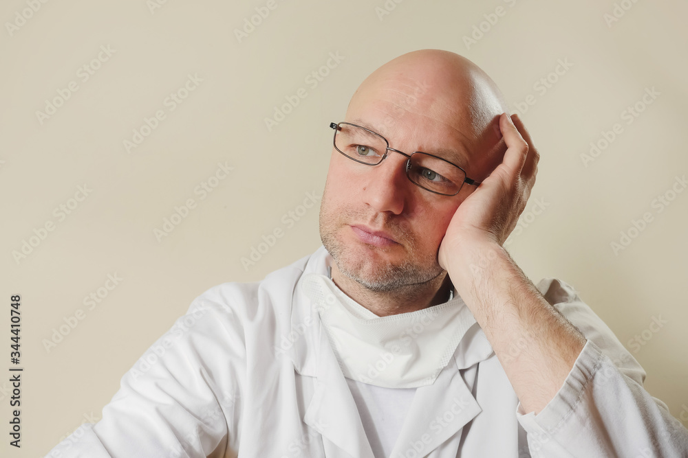 Portrait of bald unshaven doctor in metal glasses, mask under his chin, looking away. Light cream color background.