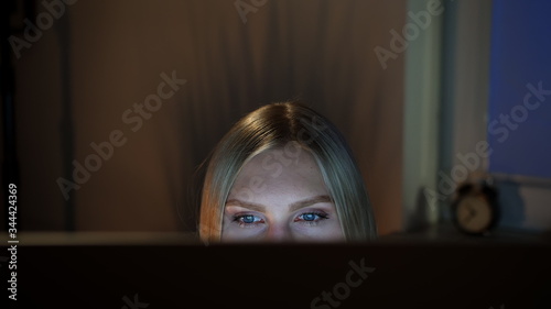Crop view of beautiful female sitting at night at computer and looking at camera over top of monitor with dark window on blurred background