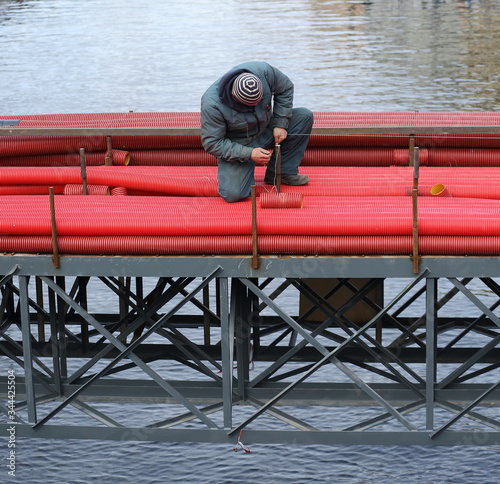 Fototapeta Naklejka Na Ścianę i Meble -  Construction worker with red pipes on a metal structure above water, Moika river, St. Petersburg, Russia, October 2019