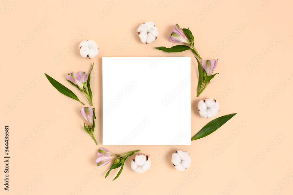 Square paper card mockup with frame made of green leaves, cotton, alstroemeria flowers on a beige background. Spring concept.