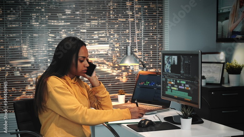 Mixed race colorist talking by smartphone and writing notes in modern production studio. In the backgrounds - skyscrapers. photo