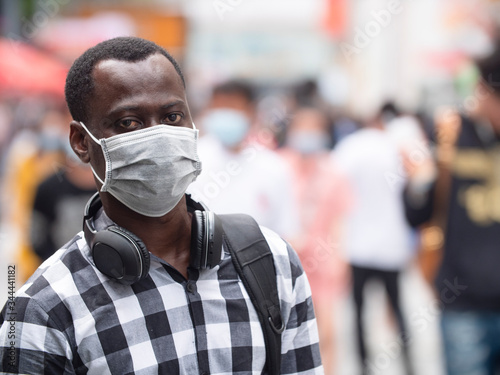  one black African man wear face mask looking at camera smile in the eye at urban street with crowd of people walking in blurred background coronavirus Covid-19 pandemic outbreak