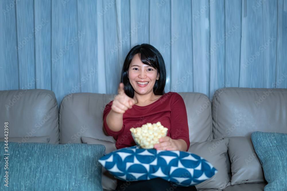 Young asian woman watching television suspense movie or news looking happy and funny and eating popcorn late night at home living room couch during time of home isolation..