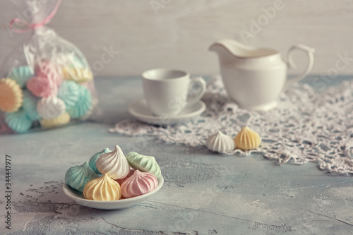 Multi-colored meringues in pastel shades with cups and a jug on a light textured wooden background. The concept of Breakfast with tea or coffee, home comfort and tenderness. Close-up, selective focus