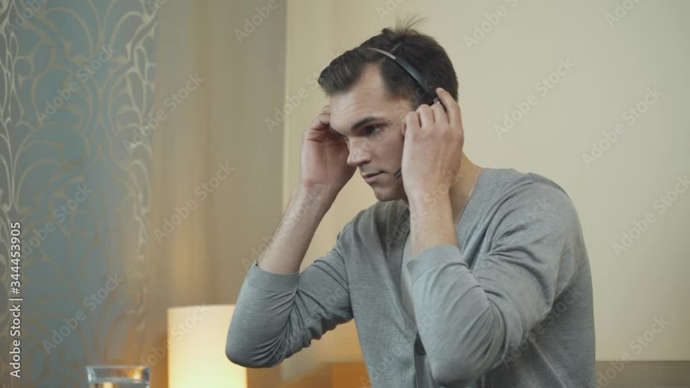 Young man puts on wireless headphone and remotely works in technical support, working at a computer and talking to the audience, work in quarantine. Young man puts on wireless headphone and remotely works in technical support, working at a computer and talking to the audience, work in quarantine.