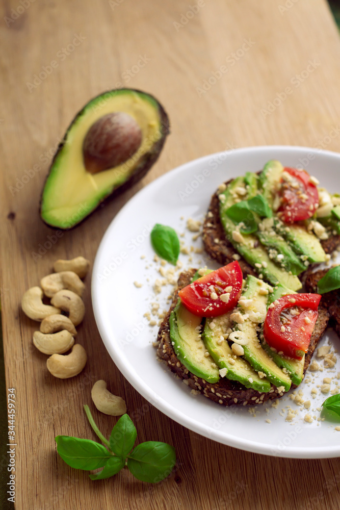Avocado sandwich on a white plate with full grain bread, tomatoes, basil and cashew nuts. Wooden background. Upright format. Avocado sandwich on a white plate with full grain bread, tomatoes, basil and cashew nuts. Wooden background. Upright format.
