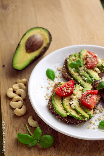 Avocado sandwich on a white plate with full grain bread, tomatoes, basil and cashew nuts. Wooden background. Upright format.