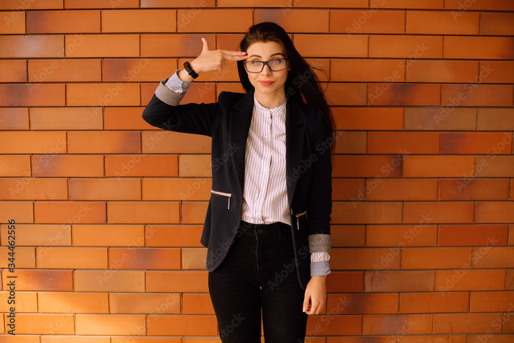 Portrait of young successful business woman with long brunette hair in glasses and suit shows gesture gun at the temple and looking at the camera, red brick wall on the background, full-length photo Portrait of young successful business woman with long brunette hair in glasses and suit shows gesture gun at the temple and looking at the camera, red brick wall on the background, full-length photo