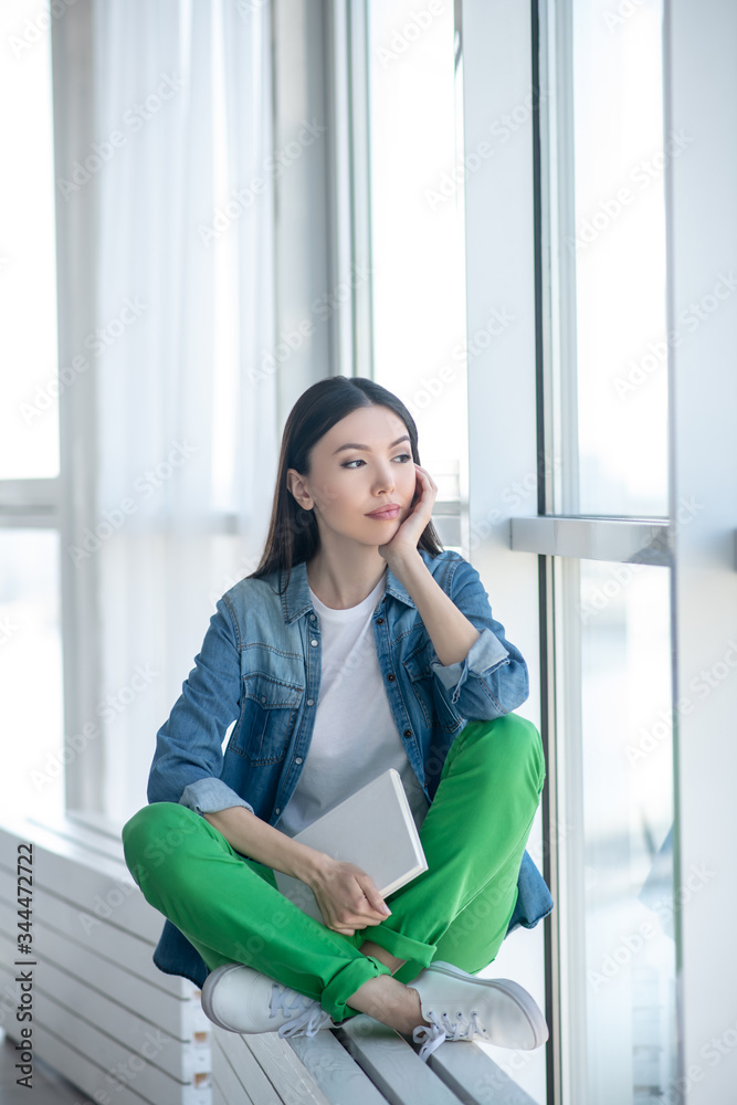 Young woman in a jeans jacket sitting near the window and looking thoughtful Young woman in a jeans jacket sitting near the window and looking thoughtful