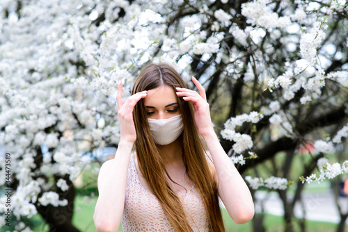 Close up portrait of tender girl in a white blouse under a blossoming cherry tree with a mask with flowers on from the coronavirus.