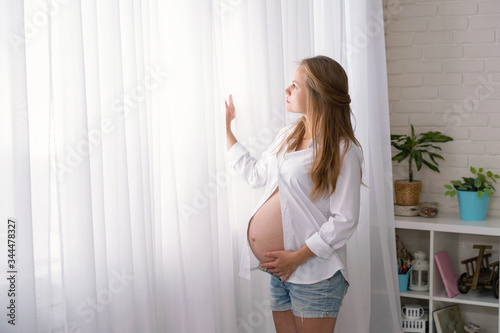 Side view portrait of a young pregnant woman, stands and looks out the window