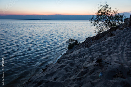 Sunset with clouds on the reservoir horizon. Sandy shore with a bush at dusk. Water with waves. Horizontal orientation.